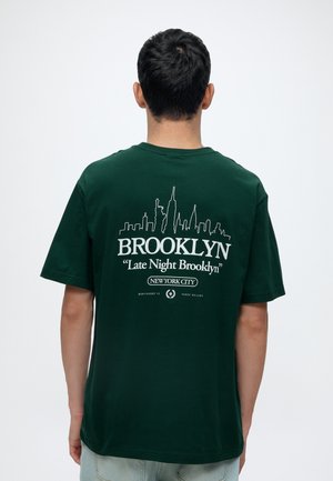 Homme portant un t-shirt vert foncé avec un graphisme blanc de la skyline de Brooklyn et le texte "Late Night Brooklyn New York City" dans le dos.