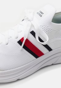 Bílé textilní tenisky s červenými a námořnickými pruhy, kulatá špička, ploché bílé tkaničky a značená nášivka na jazyku. Texturovaná podrážka.