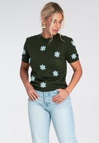 Kobieta z blond włosami nosi ciemnozielony sweter z krótkim rękawem ozdobiony jasnoniebieskimi kwiatami oraz jasnoniebieskie dżinsy, pozując z rękami na biodrach.