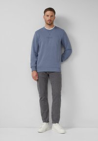 Muž v modrej mikine, šedých džínsoch a bielych teniskách, stojaci pred čistou svetlo šedou stenou.