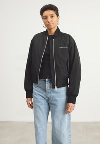 Czarna kurtka bomberka z prążkowanymi mankietami, zamkiem błyskawicznym z przodu oraz haftowanym logo "TOMMY JEANS", w połączeniu z jasnoniebieskimi dżinsami. Gładka faktura.