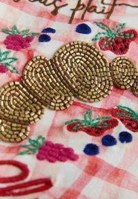 Broderet stof med en pink og hvid ternet baggrund, dekoreret med farverige frugtmotiver og guldperler arrangeret i et snoet mønster.