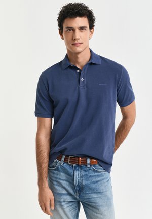 Jonge man met krullend haar, gekleed in een marineblauw poloshirt, een bruine riem en lichtblauwe jeans, staand tegen een effen witte achtergrond.