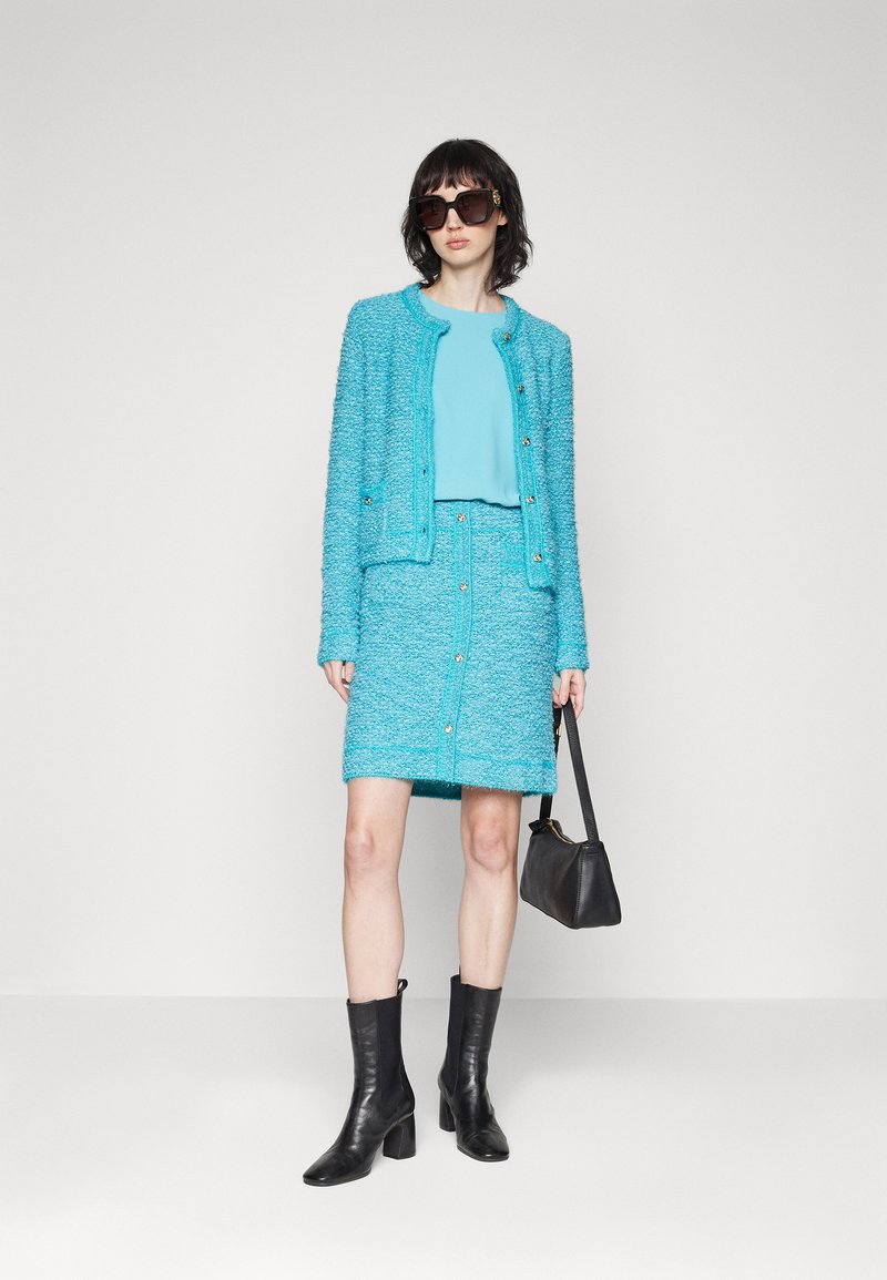 Türkiser Tweed-Blazer mit passendem Rock, mit goldenen Knöpfen. Das Model trägt ein blaues Oberteil, eine schwarze Handtasche und schwarze hohe Stiefel. Dunkle Sonnenbrille.