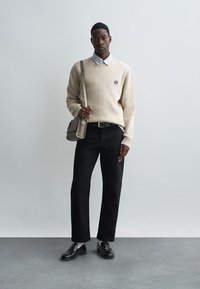 Suéter bege com um pequeno logotipo, camisa de colarinho xadrez, jeans pretos, mocassins pretos e uma bolsa transversal cinza. Fundo simples.