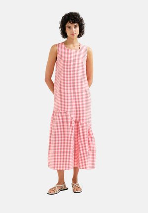 Mujer con cabello rizado y oscuro lleva un vestido midi sin mangas de cuadros rosas y blancos y sandalias blancas, de pie con las manos detrás de la espalda.