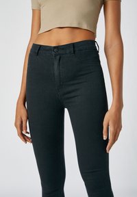 Jeans ajustados negros con una textura suave, cintura alta y detalles de costura sutiles. Modelo vistiendo un top corto de punto beige.