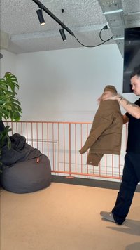 Homme tenant une veste marron dans une pièce moderne avec un fauteuil poire noir, une plante verte et des luminaires de plafond apparents.