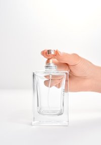 Przezroczysta szklana butelka perfum o prostokątnym kształcie, srebrna dysza rozpylająca i metalowa nakrętka trzymane w dłoni. Brak widocznego logo.
