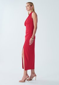 Red midi dress in stretch fabric, featuring a deep V-neck, ruched waist, and thigh-high slit. Paired with nude strappy heels.
