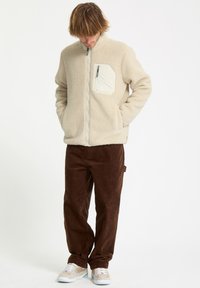 Veste en polaire crème avec poche poitrine zippée, col montant et fermeture éclair intégrale, associée à un pantalon en velours côtelé marron et des baskets.