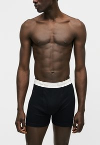 Άνδρας με σκούρο δέρμα που φοράει μαύρο boxer Calvin Klein με λευκή μέση, στέκεται μπροστά σε απλό φωτεινό φόντο.