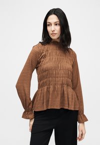 Γυναίκα με σκούρα μαλλιά που φοράει καφέ μακρυμάνικη μπλούζα με υφή και βολάν στο τελείωμα και μαύρο παντελόνι, κοιτάζοντας προς τα πλάγια μπροστά σε απλό φόντο.