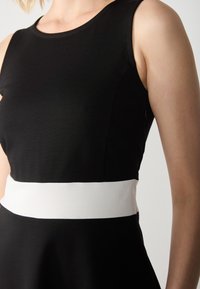 Svart ärmlös klänning med ett vitt midjeband. Slät tygkvalitet med en åtsittande bodice och en utsvängd kjol. Sidogång med dragkedja synlig.