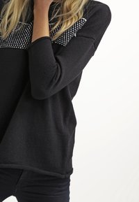 Pull en maille noir avec un design texturé, présentant un accent blanc à motif sur le haut de la poitrine et des manches trois quarts.