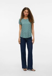 Kortærmet teal top med hvide prikker, parret med mørkeblå flagrende jeans. Modellen står på en ensfarvet lys baggrund.