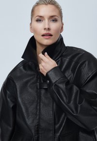 Veste en faux cuir noir avec un col haut, présentant une finition texturée, des accents de boutons et une coupe ample et surdimensionnée.