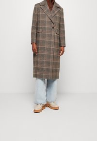 Persoon in een bruine geruite lange jas, lichtblauwe wijde jeans, beige schoenen met een dikke zool en een bruine top met een ritssluiting in de nek.