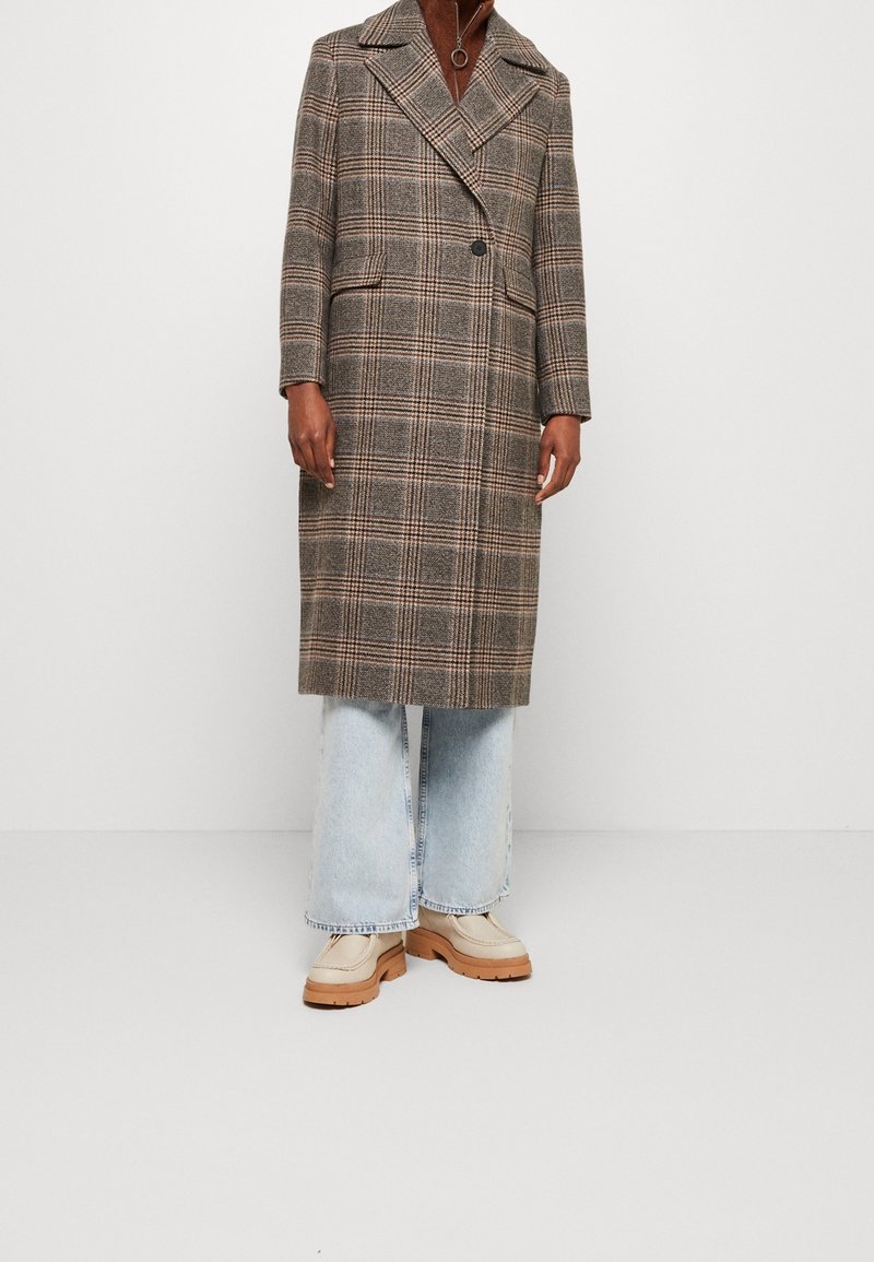 Persoon in een bruine geruite lange jas, lichtblauwe wijde jeans, beige schoenen met een dikke zool en een bruine top met een ritssluiting in de nek.