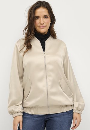 Beige satin bomber jacket with a zip front, side pockets, and elastic cuffs. Worn over a black turtleneck, featuring a smooth texture.