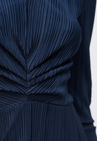 Mélykék pliszírozott ruha bemutatva. Függőleges bordázattal, testhezálló felsőrésszel, gyűrt részletekkel és bővülő szoknyával rendelkezik. Puha, texturált anyagból készült.