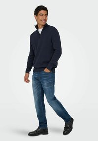 Jeune homme souriant, portant un pull à col zippé bleu marine, un jean bleu et des bottes noires, marchant avec une main dans la poche sur un fond uni.