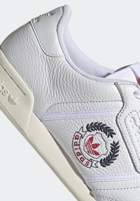 Sneaker bianco in pelle con tomaia testurizzata, dettagli cuciti, patch logo in quattro parti rosso e nero, e lacci bianchi.