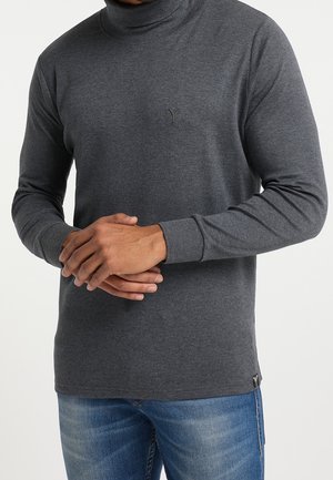 Chemise à col roulé gris à manches longues, présentant une coupe ajustée, des poignets texturés et un petit logo sur la poitrine. Portée avec un jean bleu.