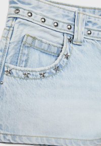 Denim bleu clair avec poche avant décorée de clous métalliques argentés en forme d'étoile et ronds le long des coutures et de la ceinture.