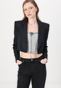 Zwarte cropped blazer with een gestructureerde afwerking, gedragen over een lichtblauwe denim top, gecombineerd met hoge zwarte jeans.