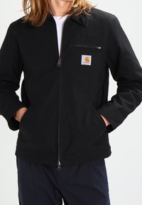 Černá zipová bunda s špičatým límcem, kapsou na prsou a bočními kapsami. Vyrobena z pevné látky, má na prsou malé logo.