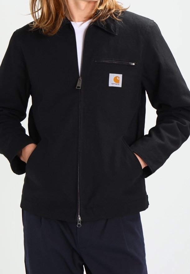 Černá zipová bunda s špičatým límcem, kapsou na prsou a bočními kapsami. Vyrobena z pevné látky, má na prsou malé logo.