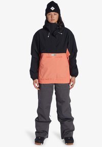 Čierna a koralová nepremokavá bunda s predným vreckom, elastickým pásom a vysokým golierom; v kombinácii so sivými nohavicami a čiernymi čižmami.