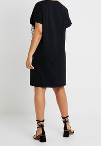 Černé bavlněné šaty s krátkými rukávy, rovného střihu a detailem na zadním švu. V doprovodu černých sandálů na podpatku s blokovým podpatkem.