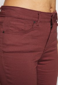 Jeans burdeos hechos de denim elástico, con un bolsillo frontal, costuras sutiles y una cintura de altura media con lazos para el cinturón.