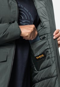 Grüne isolierte Jacke mit Reißverschluss, ausgestattet mit einer Seitenasche zur Aufbewahrung und einem sichtbaren Etikett mit der Aufschrift "Texapore Ecosphere."