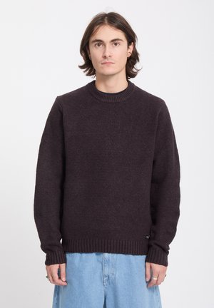Jeune personne aux cheveux bruns mi-longs, portant un pull marron foncé et un jean bleu clair, faisant face à l’avant sur un fond uni.