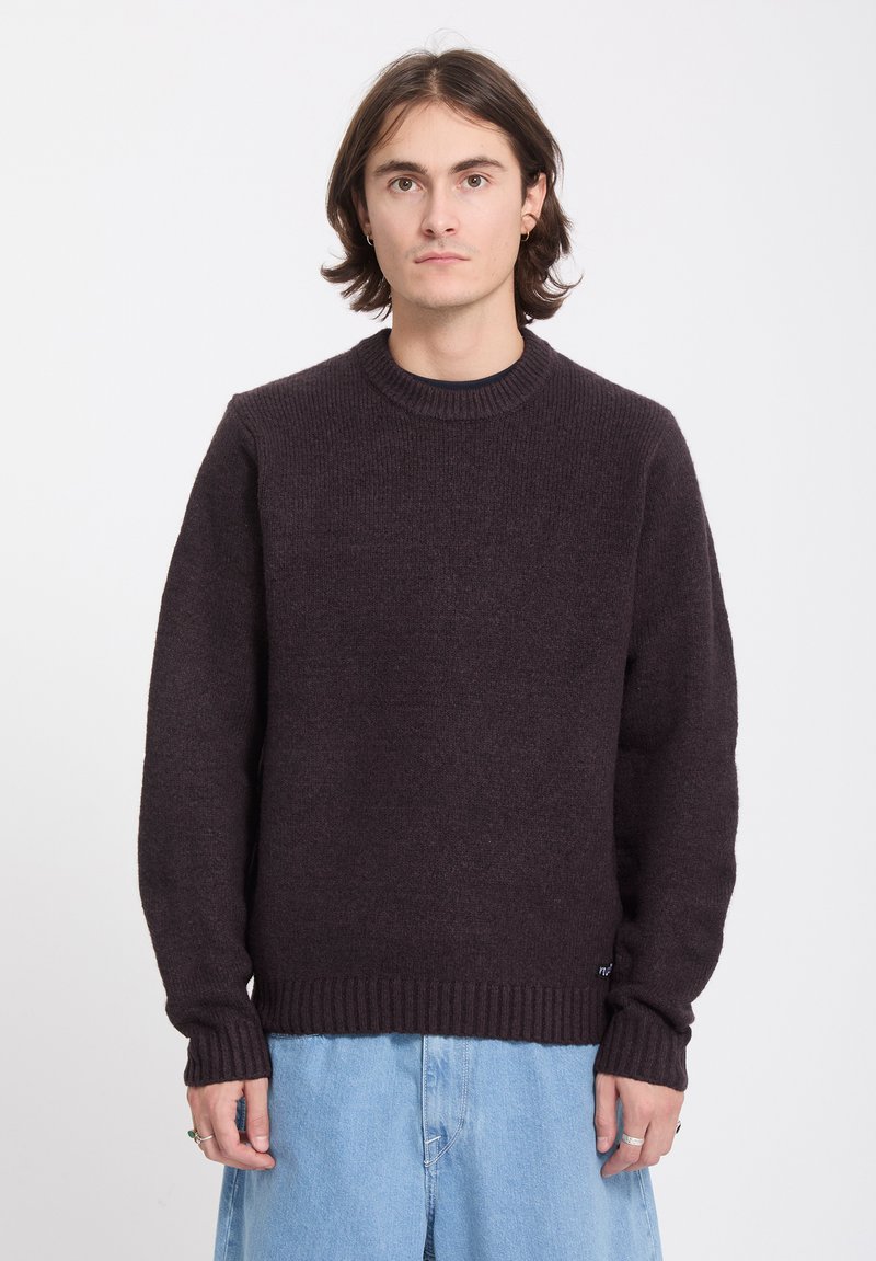 Jeune personne aux cheveux bruns mi-longs, portant un pull marron foncé et un jean bleu clair, faisant face à l’avant sur un fond uni.
