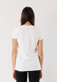 T-shirt bianco in cotone a maniche corte, collo a giro, con un discreto etichetta rosa sul retro. Indossato con pantaloni neri aderenti.