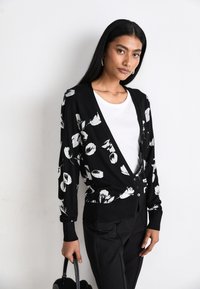Svart stickad cardigan med vitt blommönster, v-ringning, långa ärmar och framknapp. Stylist med en vit topp och svarta byxor.