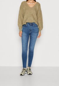 Maglione lavorato a maglia di colore khaki con scollatura a V, abbinato a jeans skinny blu a vita alta e sneakers beige con dettagli neri e lacci.