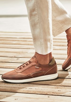 Zapatillas de cuero marrón con paneles de malla, usadas con pantalones beige claro sobre suelo de madera.