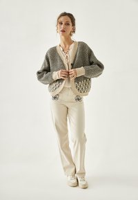 Cárdigan de punto con patrón de espiga en negro y crema, cierre de botones, combinado con pantalones beige con bordado floral. Zapatillas blancas.