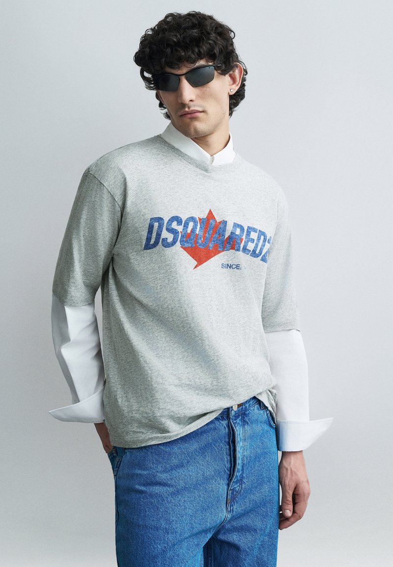 Graues Baumwoll-T-Shirt mit rotem und blauem "DSQUARED"-Logo und Ahornblatt-Grafik. Darüber ein weißes Hemd mit Knöpfen, kombiniert mit blauen Jeans.
