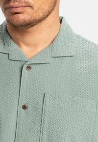 Light green short-sleeve shirt with a textured fabric, featuring a collar, a chest pocket, and a brown button. Pattern is subtly woven.