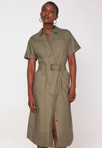 Donna che indossa un vestito camicia verde oliva con cintura, maniche corte e bottoni frontali, in piedi su uno sfondo bianco semplice.