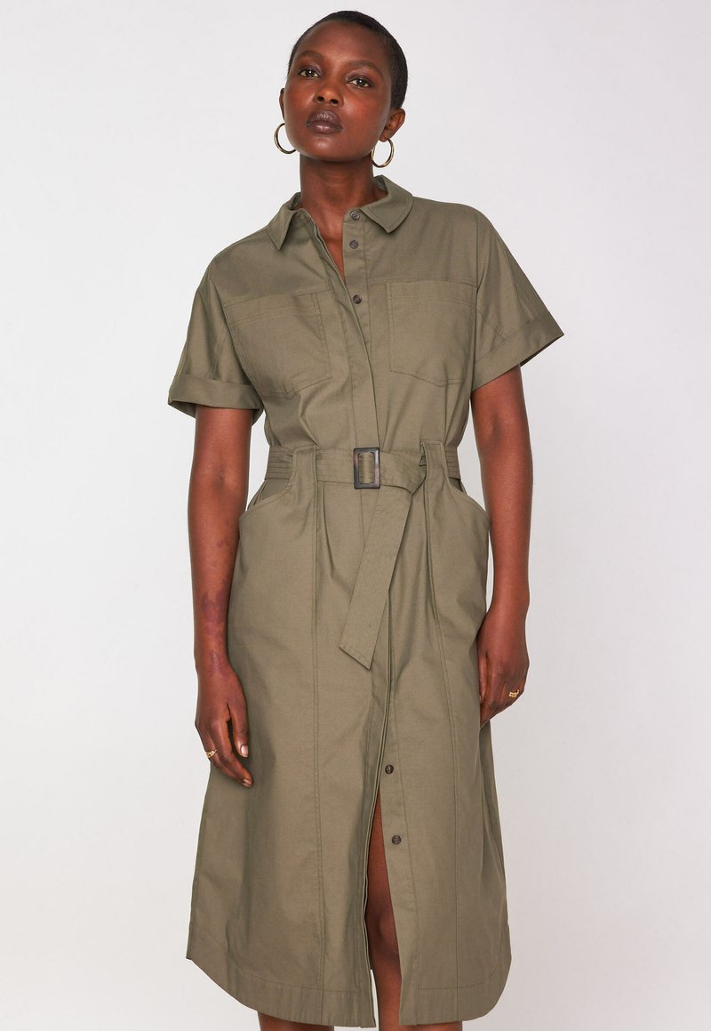 Donna che indossa un vestito camicia verde oliva con cintura, maniche corte e bottoni frontali, in piedi su uno sfondo bianco semplice.