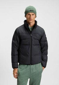 Black puffer jacket with a high collar, zip closure, and horizontal quilted pattern. Paired with a green beanie featuring 'BOSS' logo.