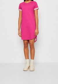 Różowa bawełniana sukienka z krótkimi rękawami, okrągłym dekoltem, bocznymi rozporkami i kontrastującymi jasnoróżowymi mankietami. Noszona z beżowymi botkami za kostkę.