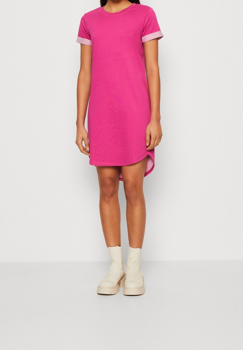 Różowa bawełniana sukienka z krótkimi rękawami, okrągłym dekoltem, bocznymi rozporkami i kontrastującymi jasnoróżowymi mankietami. Noszona z beżowymi botkami za kostkę.